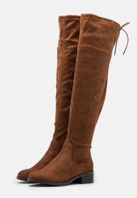 Bottes en suède marron à hauteur de genou avec un bout arrondi et un talon carré. Présentent un tige flexible et un lien réglable en haut.