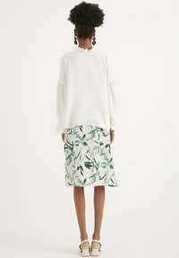Hvid bluse med flæsekrave, parret med en knælang nederdel med et grønt botanisk print. Enkle sandaler fuldender looket.