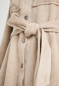 Robe en velours côtelé beige avec ceinture nouée, texture côtelée verticale, boutons argentés et poches plaquées sur la poitrine.