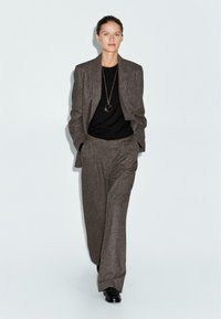 Completo grigio in lana con giacca su misura e pantaloni a gamba larga, abbinato a un maglione nero e una lunga collana con pendente. Scarpe nere.