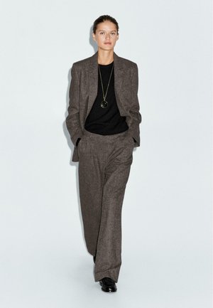 Completo grigio in lana con giacca su misura e pantaloni a gamba larga, abbinato a un maglione nero e una lunga collana con pendente. Scarpe nere.
