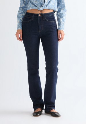 Kvinde iført mørkeblå jeans med lige ben, sorte balletsko og en lyseblå blomstret langærmet skjorte bundet i taljen.