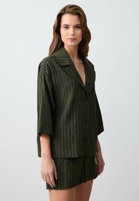 Junge Frau in einer dunkelgrünen, strukturierten Knopfjacket und passenden Shorts, die vor einem schlichten hellen Hintergrund steht.