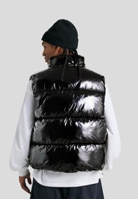Crni puffer prsluk, sjajan, s kvalitetnom qiltanom teksturom i visokim ovratnikom. Ispod njega bijela košulja s dugim rukavima. Pogled s leđa, ležeran stil, minimalistički dizajn.