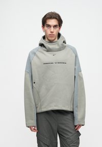 Nike Tech πουλόβερ σε ανοιχτό γκρι χρώμα με υφασμάτινους μπλε ώμους, διαθέτει ψηλό λαιμό, μαύρο λογότυπο και λεπτομέρεια πλευρικών αεραγωγών.