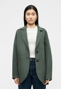 Γυναίκα με μακριά σκούρα μαλλιά που φοράει χρυσούς κρίκους στα αυτιά, πράσινο oversized σακάκι, λευκή ριμπ μπλούζα και σκούρο μπλε τζιν, στέκεται μπροστά σε λευκό φόντο.