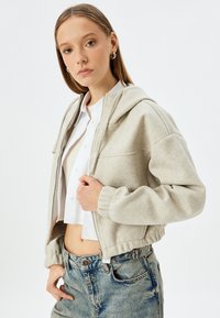 Helle graue, kurz geschnittene Jacke aus weichem Fleece-Material mit Kapuze, gerippten Bündchen und einem Reißverschluss vorne, kombiniert mit einem weißen Hemd mit Kragen und Jeans.