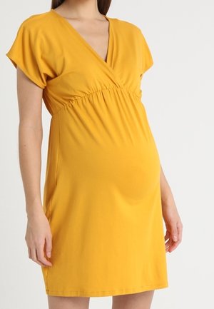 Femme enceinte portant une robe de maternité à manches courtes, jaune moutarde, col en V avec un détail froncé à la taille.