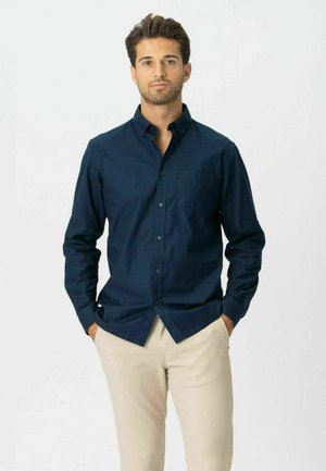 Mężczyzna o ciemnych włosach, ubrany w granatową koszulę na guziki i beżowe spodnie, stoi z rękami w kieszeniach na tle jednolitego białego tła.