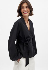 Blusa negra envuelta con mangas largas, que cuenta con un cinturón atado en la cintura y un diseño de cuello en V. La tela texturizada añade profundidad al conjunto.