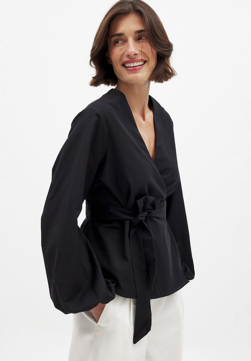 Blusa negra envuelta con mangas largas, que cuenta con un cinturón atado en la cintura y un diseño de cuello en V. La tela texturizada añade profundidad al conjunto.
