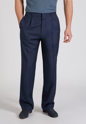 Mężczyzna w granatowych, dopasowanych spodniach i ciemnoniebieskich mokasynach, ręce swobodnie opuszczone wzdłuż ciała, stojący na jasnoszarej podłodze.