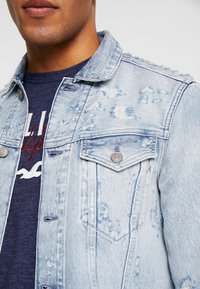 Light blue denim jacket featuring distressed details, two chest pockets, metal buttons, and a classic collar design. Worn over a navy t-shirt.