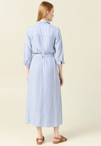 Abito lungo con bottoni di colore azzurro chiaro a strisce bianche. Presenta una vita con cintura, maniche a tre quarti e colletto. Indossato con sandali marroni.