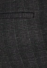 Tissu gris foncé avec un motif à carreaux discret et une couture horizontale de poche passepoilée, montrant la texture et les détails finement tissés.