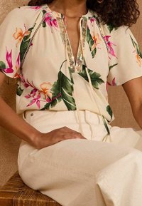 Blumige Bluse in Creme mit rosa und grünen Akzenten, ausgestattet mit einem V-Ausschnitt, der mit goldenen Pailletten verziert ist, und einer Schleifendetail. Kombiniert mit beigen Hosen.