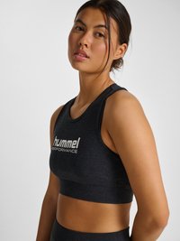 Jonge vrouw met donker haar naar achteren gebonden, die een zwarte mouwloze cropped top draagt met het "hummel PERFORMANCE" logo, poserend tegen een effen lichte achtergrond.