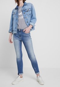 Džínová bunda ve světle modré barvě s knoflíkovým zapínáním a náprsními kapsami, doplněná pruhovanou šedou košilí a světle modrými skinny džíny, dokončená bílými teniskami.