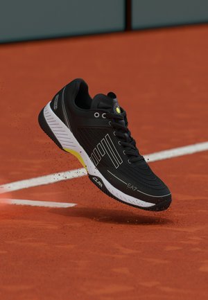 Sort-hvid tennissko, der sparker op lerstøv på en rød lerbane nær en hvid linje.