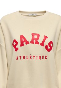 Beige svetr s kulatým výstřihem, který má výrazný červený text "PARIS" a "ATHLÉTIQUE" ve zakřiveném designu. Měkký bavlněný materiál, volný střih.