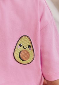 Roosa T-särk tekstuurse tikitud avokaado kujutisega ees, millel on rohelised ja pruunid toonid, naeratav nägu ja punetavad põsed.