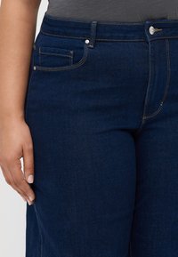 Mørkeblå denimjeans med en glatt tekstur, en frontlomme og sølvfarget spenne i midjen. Syde detaljer langs sømmene.