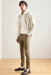 Gestreept overhemd met knopen in groen en wit, gedragen over een lichtcrèmekleurig shirt. Lichtbruine broek en bruine loafers maken de outfit compleet.