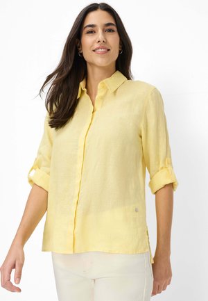 Vrouw met lang donker haar, rolmouwen opgerold, gele blouse met knopen en lichtgekleurde broek, licht glimlachend, tegen een witte achtergrond.