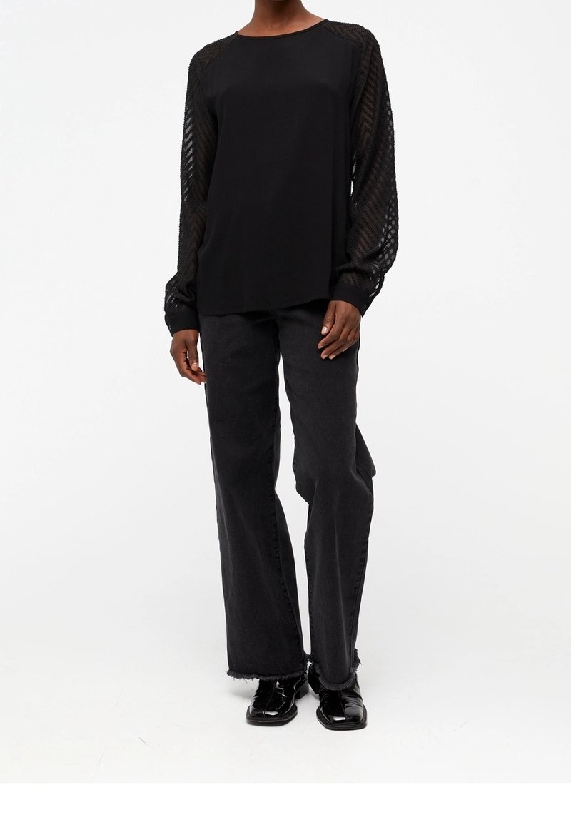 Haut noir avec corps uni et manches en tissu transparent à motifs, accompagné d'un pantalon noir taille haute et de chaussures noires brillantes. Design simple et moderne.