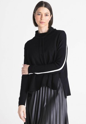 Vrouw met steil donker haar draagt een zwarte trui met witte mouwstrepen en een geplooide donkergrijze rok, staand tegen een witte achtergrond.