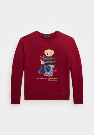 Czerwona bluza z długim rękawem z misiem w swetrze i dżinsach, trzymającym zapakowany prezent oraz napisem "POLO BEAR by RALPH LAUREN."
