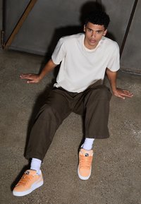Light beige t-shirt, brown wide-leg pants, and orange suede sneakers with white sole and laces. Texture is smooth, with visible stitching details.