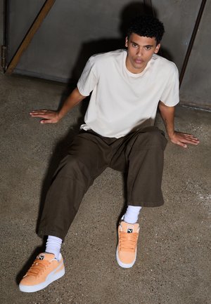 Lichtbeige t-shirt, bruine wijde broek en oranje suède sneakers met een witte zool en veters. De textuur is glad, met zichtbare stikseldetails.