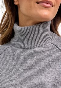 Grijze gebreide coltrui met geribbelde textuur, voorzien van een hoge kraag en een licht ontspannen pasvorm. Close-up toont zachte stofdetails.