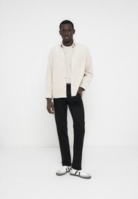 Világos bézs gombos ing világos felső fölött, fekete nadrággal és fehér sportcipőkkel, amelyeken szürke és fekete díszítések vannak.