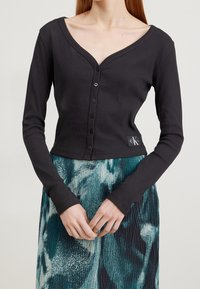 Svart ribbad långärmad cardigan med djup V-ringning, med knappstängning och en märkesskylt vid fållen, i kombination med en mönstrad kjol.