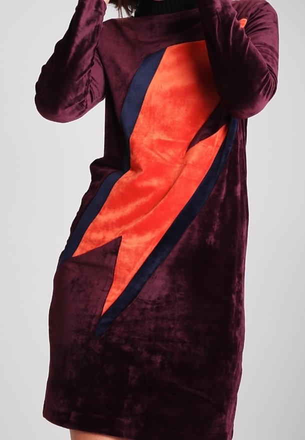 Femme portant une robe en velours violet avec un grand motif d’éclair orange et noir sur le devant.