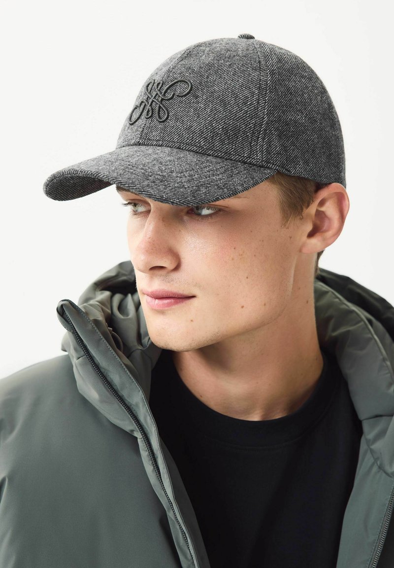 Grå uldblanding baseball cap med syet logo, buet skygge og syet detaljer. Sammen med en grå jakke med lynlås.