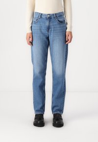 Vaqueros de mezclilla azul claro con un diseño de pierna recta, cinco bolsillos y cierre de botón. Usados con una blusa crema y zapatos negros.