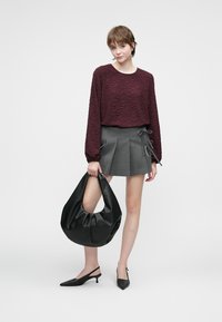 Pullover testurizzato color bordeaux, mini gonna pieghettata grigia con laccetti laterali, borsa hobo nera e décolleté con punta a stiletto neri su uno sfondo neutro.