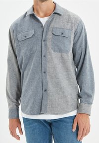 Grau gestreiftes Hemd mit Knöpfen, zwei Brusttaschen, langen Ärmeln und einem strukturierten Stoff, kombiniert mit einem weißen Unterhemd und blauen Jeans.