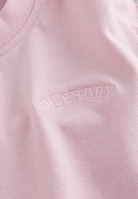 Tissu rose pâle avec texte brodé ton sur ton "SUPERMAY" en lettres majuscules courbes près de la couture.