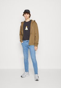 Sudadera marrón con cremallera sobre una camiseta negra con un gráfico colorido, jeans de mezclilla azul claro y zapatillas grises con detalles negros. Gorra negra.