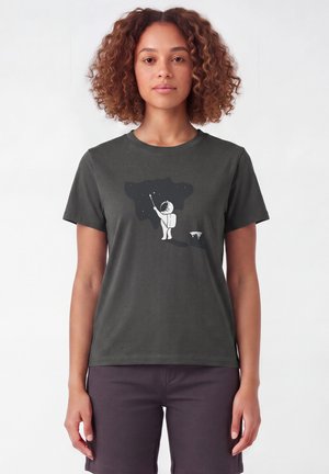 Femme aux cheveux bouclés portant un t-shirt gris foncé montrant un astronaute peignant des étoiles sur une forme de ciel noir.