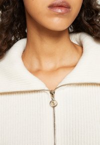 Weißes, mit Reißverschluss versehenes Kleidungsstück mit einer gerippten Struktur und einem breiten Kragen. Goldener Reißverschluss und Zipper-Akzente, Fokus auf den Hals- und Kragenbereich.