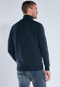 Dunkelblauer gestrickter Pullover mit geripptem Kragen und horizontalem Streifenmuster. Verfügt über lange Ärmel und eine enganliegende Passform. Wird mit blauen Jeans getragen.