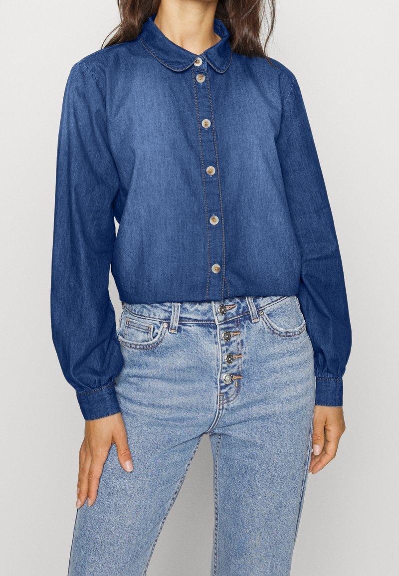 Mörk denim skjorta med krage, långa puffärmar och knappstängning; tillsammans med ljusblå high waist jeans med dekorativa knappar.