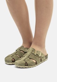 Sabots en daim vert olive avec un design à bout ouvert, des détails de sangle croisée, une boucle sur le côté et une semelle en caoutchouc beige texturé.