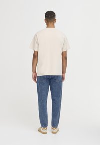 Homme debout, vu de dos, portant une chemise beige à manches courtes, un jean bleu et des baskets Adidas beiges sur un fond blanc.