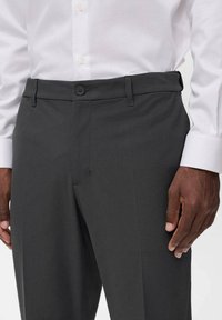 Pantaloni grigio scuro con una texture liscia, chiusura a bottone e vestibilità classica, abbinati a una camicia bianca con colletto.